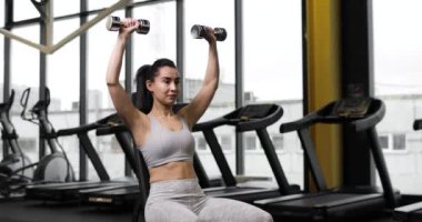 Genç esmer kadın dambıllarla üstten baskı yapıyor, spor salonundaki bir bankta oturuyor. Arka planda büyük bir pencere var..