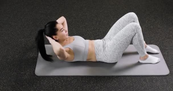 A young caucasian athletic woman does an exercise on the abdominal press, lies on the floor in the gym, dressed in gray sportswear. Side view. — 图库视频影像