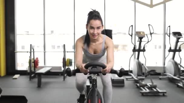 Portrait of a beautiful young brunette woman doing cardio on a exercise bike at gym. Moving away video. — стоковое видео