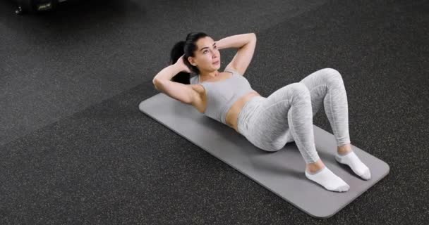 A young athletic woman does an exercise on the abdominal press, lies on the floor in the gym, dressed in gray sportswear. Top view. — Stockvideo