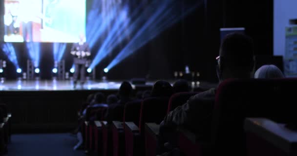 Back view between the rows of the audience sitting at a business conference. Subdued light in the hall. Speaker is holding a presentation to business people. — Vídeo de Stock