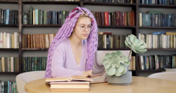 Νεαρή γυναίκα με μακριά χρωματιστά μαλλιά κάθεται σε ένα γραφείο με ένα φορητό υπολογιστή, που εργάζεται στη βιβλιοθήκη. — Αρχείο Βίντεο