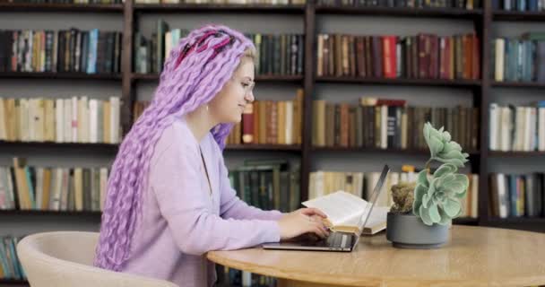 Молодая женщина с длинными волосами сидит за столом с ноутбуком, работая в библиотеке. Вид сбоку. — стоковое видео
