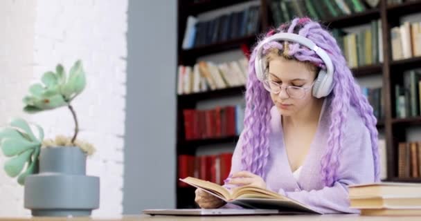 Una mujer joven en auriculares con el pelo largo de color se sienta en una mesa con un ordenador portátil y una planta de interior, trabaja en la biblioteca. Movimiento lento de la cámara. — Vídeos de Stock