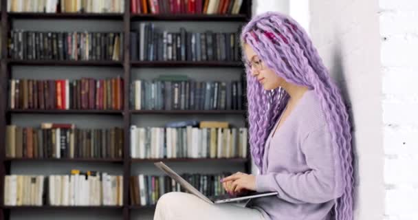 Jovem com cabelos longos e coloridos está trabalhando sentado na biblioteca com um laptop. Vista lateral. — Vídeo de Stock