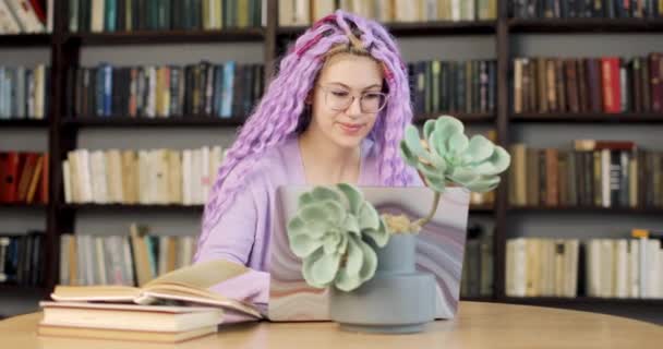 Young woman with long colored hair is sitting at a desk with a laptop, working in the library. Front view. Slow camera movement. — Stock Video