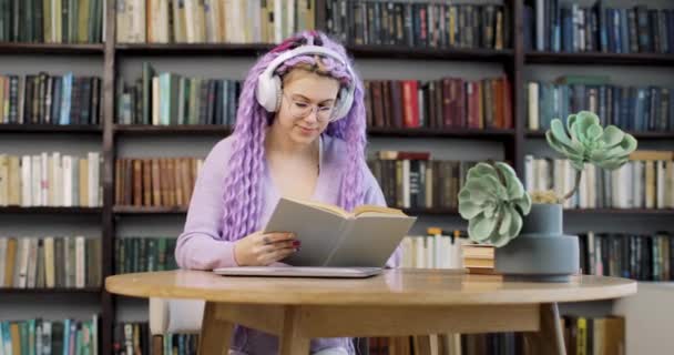 Uma jovem mulher em fones de ouvido com cabelos longos e coloridos senta-se em uma mesa com um laptop e planta de sala, trabalha na biblioteca. Vista frontal. — Vídeo de Stock