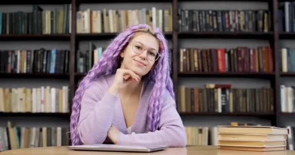 Retrato de una mujer joven con el pelo largo de color púrpura sentado en un escritorio con un ordenador portátil y libros, en la biblioteca. Sonriendo y mirando a la cámara. Vista frontal. — Vídeos de Stock