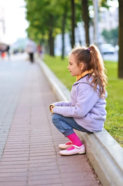 Little girl in jacket sitting on paving-stone curb Stock Photo