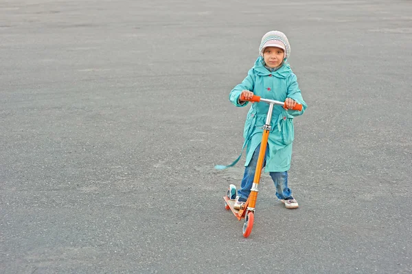 Malá holčička v modrém plášti jezdecké skútr — Stock fotografie