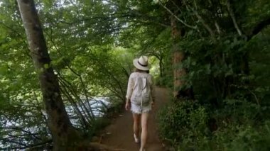 Şapkalı genç bir kadın göl boyunca yürüyor.