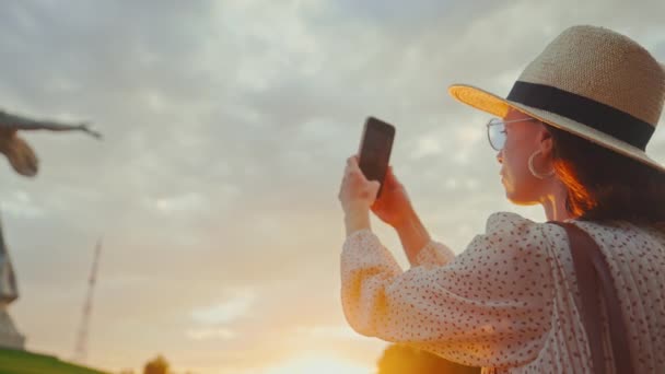 Μια ελκυστική γυναίκα κοντά στη Μητέρα Πατρίδα τραβώντας μια φωτογραφία σε ένα smartphone στο ηλιοβασίλεμα — Αρχείο Βίντεο