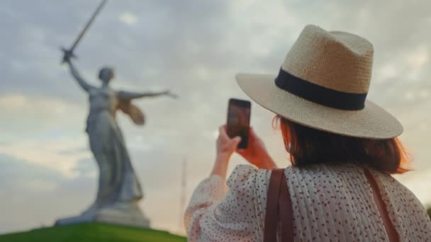 En attraktiv kvinde i en hat nær Motherland tager et billede på en smartphone på Mamayev Kurgan. Volgograd, Rusland – Stock-video