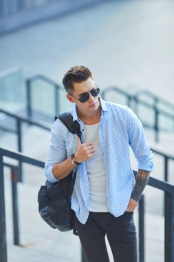 Siyah sırt çantalı, güneş gözlüklü genç bir adam merdivenlerde durup korkuluklara yaslanıyor..