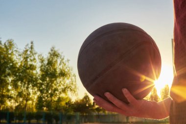 Profesyonel basketbolcunun elinde top tutmasına yakın çekim. Arka planda mavi gökyüzü ve top ile sokak basketbolu atleti