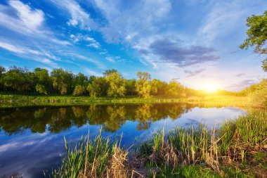 Yazın gün batımında güzel nehir kıyısında. Göl, yeşil ağaçlar ve çimenler ile renkli manzara, mavi gökyüzü çok renkli bulutlar ve suya yansıyan turuncu güneş ışığı. Doğa. Canlı bir manzara.