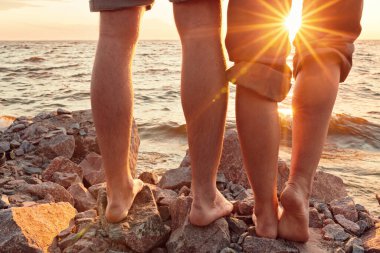 Bacakları sahilde. Ayak spası. Gün batımında sahilde sarılıp öpüşen genç bir çift. İki sevgili, erkek ve kadın, suyun yanında yalınayak. Yaz aşkı.