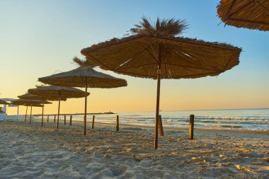 Deniz kıyısı, şemsiyeli kumsal ve şafakta deniz ve gökyüzünün arka planında güneşlik.