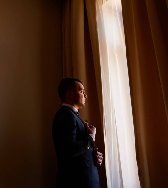 Beyaz gömlekli genç adam pencerenin yanındaki kol düğmelerini takıyor. İş portresi. Damat gelinle buluşacak. Düğün için. İş adamı. Nişanlı.