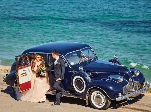 Счастливая невеста и жених обнимаются и позируют возле старого ретро-автомобиля перед свадебной церемонией