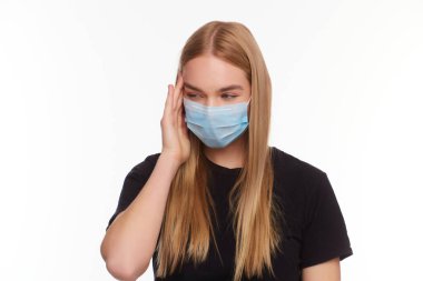 Kapalı gözlü, tıbbi grip maskesi takan, başını eliyle tutarak beyaz arka planda izole edilmiş genç sarışın kızın stüdyo portresi..