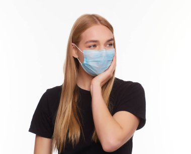 Kadın virüse karşı koruyucu beyaz bir maske takıyor, şakaklarında şiddetli bir acı hissediyor, parmaklarını kafaya doğru tutuyor. Siyah tişörtlü, uzun boylu sarışın bir kız. Stüdyoda beyaz arka planda izole edilmiş.