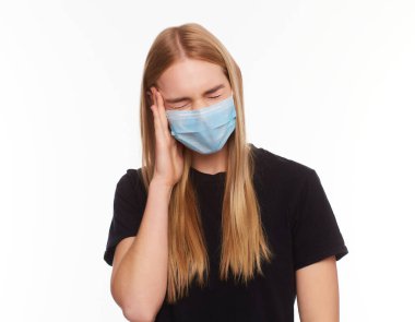Kadın virüse karşı koruyucu beyaz bir maske takıyor, şakaklarında şiddetli bir acı hissediyor, parmaklarını kafaya doğru tutuyor. Siyah tişörtlü, uzun boylu sarışın bir kız. Stüdyoda beyaz arka planda izole edilmiş.