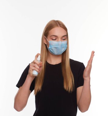 Yüzünde koruyucu bir tıbbi maske takan genç bir kız, izole edilmiş beyaz bir arka planda bir dezenfektan, el antiseptiği tutuyor. Virüs kavramı, koronavirüs..