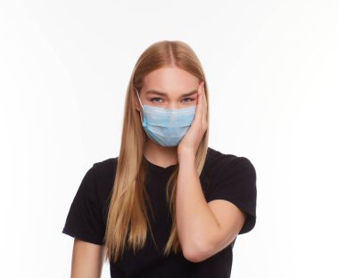 Kapalı gözlü, tıbbi grip maskesi takan, başını eliyle tutarak beyaz arka planda izole edilmiş genç sarışın kızın stüdyo portresi..