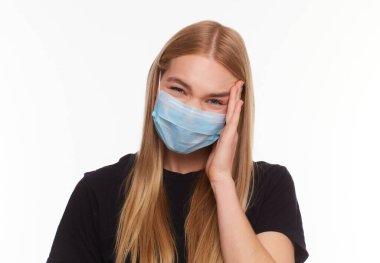 Kapalı gözlü, tıbbi grip maskesi takan, başını eliyle tutarak beyaz arka planda izole edilmiş genç sarışın kızın stüdyo portresi..