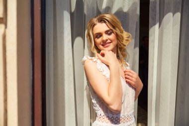 Gelin sabahı hazırlığı. Beyaz gelinlik içinde güzel bir gelin ve büyük yatak odası penceresinin arkasında peçe. Gelinin ücreti. Gelin gelinliğini giymek üzere.