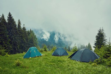 Çimenli tepede, sisli dağların üzerindeki güzel mavi gökyüzünün altında orman zeminiyle kaplı küçük, rahat bir turist çadırının yaz manzarası. Kamp, turizm ve seyahat konsepti.