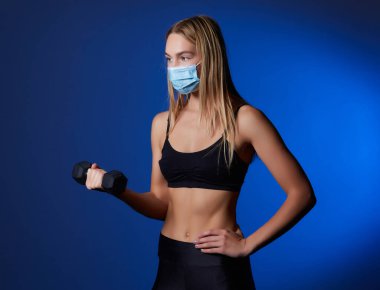 Coronavirus 'un yayılmasını önlemek için maske takan sağlıklı bir kadın halterle biseps kıvırıyor. sağlıklı yaşam tarzı ve spor konsepti.