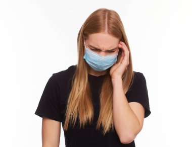 Kadın virüse karşı koruyucu beyaz bir maske takıyor, şakaklarında şiddetli bir acı hissediyor, parmaklarını kafaya doğru tutuyor. Siyah tişörtlü, uzun boylu sarışın bir kız. Stüdyoda beyaz arka planda izole edilmiş.