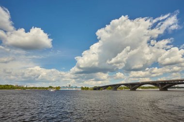 Dinyeper Nehri 'nin sağ kıyısından Kyiv şehrindeki ilk metro köprüsünün üzerinden mavi metro treni, altında motorlu tekne ve arkasında otellik görünüyor. Mavi gökyüzüyle güneşli bir gün.