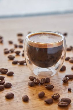 Arkaplanda kahve çekirdekleri ile çevrili, ahşap bir masanın üzerinde duran şeffaf bir bardak sıcak espresso.