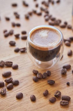 Şeffaf bir fincanda Espresso, ahşap bir masanın üzerinde duruyor, arka planda kahve çekirdekleri ile çevrili.