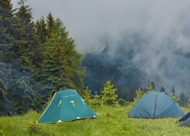 Çimenli tepede, sisli dağların üzerindeki güzel mavi gökyüzünün altında orman zeminiyle kaplı küçük, rahat bir turist çadırının yaz manzarası. Kamp, turizm ve seyahat konsepti.