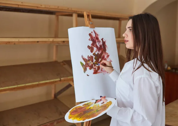 白いシャツを着た若いブルネットのアーティストが彼女のスタジオでキャンバスにペイントします 彼女の隣にはイーゼル 様々な芸術技術があります — ストック写真