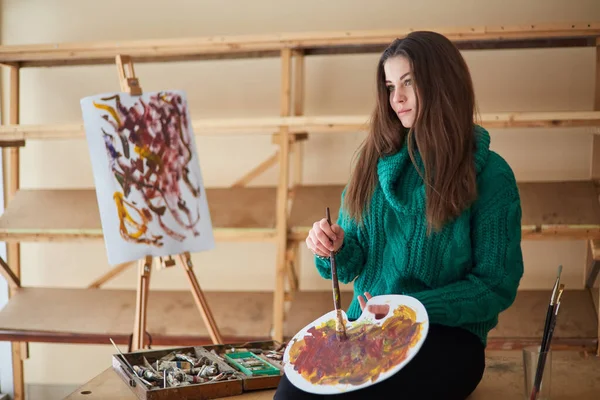 The girl artist, brunette, paints a colorful picture. Mixes paints on a palette, a close-up of a painting process in an art workshop. A creative positive woman painter paints an abstract picture in her workshop.