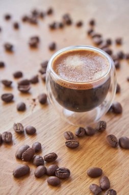 Şeffaf bir fincanda Espresso, ahşap bir masanın üzerinde duruyor, arka planda kahve çekirdekleri ile çevrili.