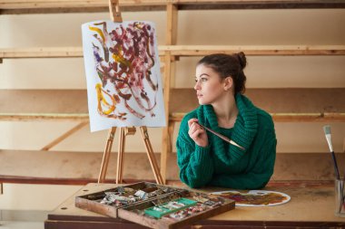 Esmer sanatçı kız elinde fırçalarla sehpanın yanında duruyor. Sanat atölyesinde yaratıcı bir ortam