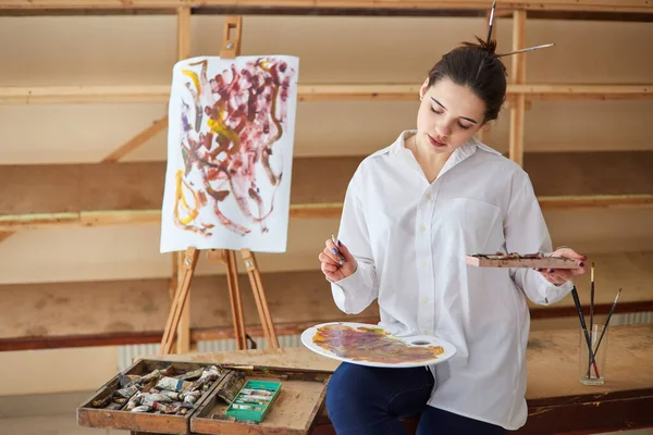女の子の芸術家 ブルネットは カラフルな絵を描きます パレットに絵具を混ぜ アートワークショップで絵具のプロセスを閉じます 創造的な肯定的な女性画家は 彼女のワークショップで抽象的な絵を描きます — ストック写真