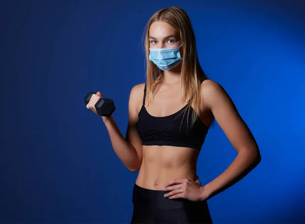 A fit woman in a face mask to avoid the spread of coronavirus is doing bicep curls with dumbbells. healthy lifestyle and sport concept.