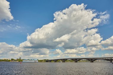 Dinyeper Nehri 'nin sağ kıyısından Kyiv şehrindeki ilk metro köprüsünün üzerinden mavi metro treni, altında motorlu tekne ve arkasında otellik görünüyor. Mavi gökyüzüyle güneşli bir gün.