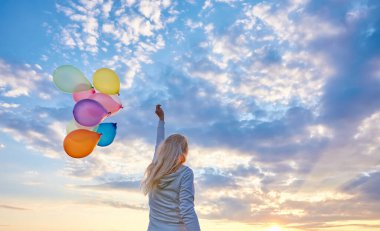 Gün batımında çayırlarda renkli balonlarla koşan genç bir kadın.