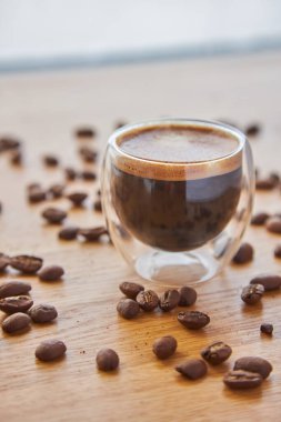 Arkaplanda kahve çekirdekleri ile çevrili, ahşap bir masanın üzerinde duran şeffaf bir bardak sıcak espresso.