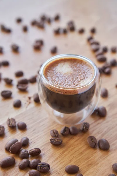 Şeffaf bir fincanda Espresso, ahşap bir masanın üzerinde duruyor, arka planda kahve çekirdekleri ile çevrili.
