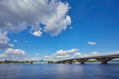Dinyeper Nehri 'nin sağ kıyısından Kyiv şehrindeki ilk metro köprüsünün üzerinden mavi metro treni, altında motorlu tekne ve arkasında otellik görünüyor. Mavi gökyüzüyle güneşli bir gün.