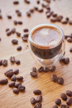 Arkaplanda kahve çekirdekleri ile çevrili, ahşap bir masanın üzerinde duran şeffaf bir bardak sıcak espresso.